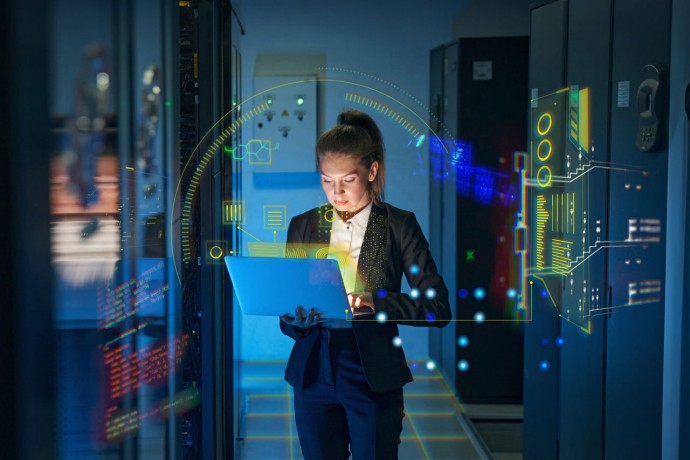 Young woman, IT engineer, uses laptop computer while working with supercomputer in data center server room